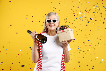 Excited senior woman with gift and bottle of sparkling wine on yellow background with flying...
