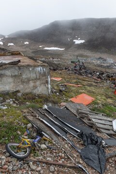 Broken children's bicycle, pallets, plywood, junkyard in rocky, hilly landscape with snow remains, arctic settlement Ittoqqortoormiit, Scoresbysund or Scoresby Sund or Greenlandic Kangertittivaq, East Greenland, Greenland