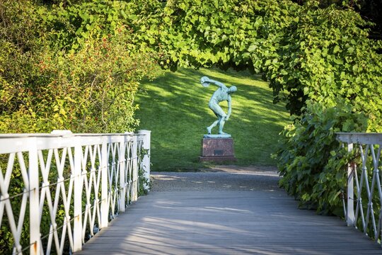 Wooden bridge leading to the sculpture of a discus launcher in a green, sunny park area, Botanical Garden or Botanisk Have, University of Copenhagen, Denmark