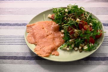 Top-down view of salmon slices with arugula, cherry tomatoes, cheese, pumpkin seeds, and oil on an olive-colored plate. Fresh, healthy, and perfect for a cozy meal.