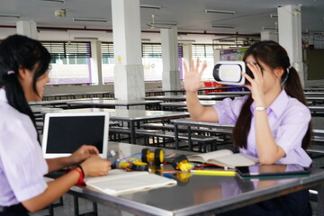 teenage female student in her school uniform sits in class doing homework and working on a robot circuit with her female friend wearing VR glasses in the cafeteria at a high school.