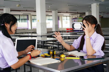  teenage female student in her school uniform sits in class doing homework and working on a robot circuit with her female friend wearing VR glasses in the cafeteria at a high school.