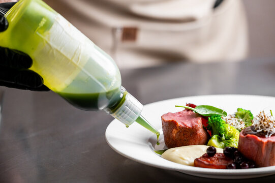 A chef in black gloves carefully garnishes a beautifully plated gourmet dish with a green sauce from a squeeze bottle. The focus is on the intricate process of fine dining presentation.