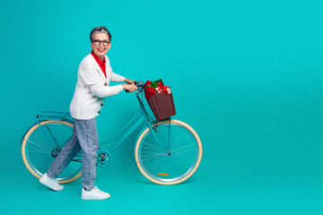 Elderly woman smiles while riding a teal bicycle with a gift basket during Christmas season against...