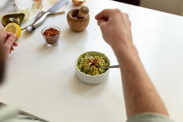 POV guacamole preparation: hand sprinkling spices on bowl. Lemon, chili, wooden mortar on table. Authentic home cooking. Fresh natural culinary aromatic traditional Mexican atmosphere.