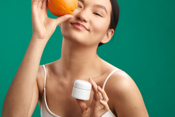asian woman holding an orange near her eye and a small skincare jar in a bright studio, radiating...
