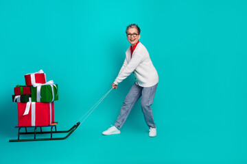 Elderly woman in a cozy cardigan pulls a sled loaded with Christmas presents against a teal studio...