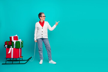 Mature woman with glasses pulls a sled stacked with gifts in a bright turquoise studio celebrating...