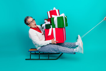 Elegant elderly woman in a white cardigan sits on a sled balancing a tall pile of red and green...