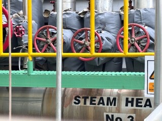 Red handwheels on industrial steam heat valves behind a yellow railing, showing insulated pipes and the "STEAM HEAT No. 3" label, symbolizing energy and heavy industry.