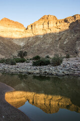 The sandstone cliffs of the Fish River Canyon reflecting into the river in its base in the golden light of the morning.
