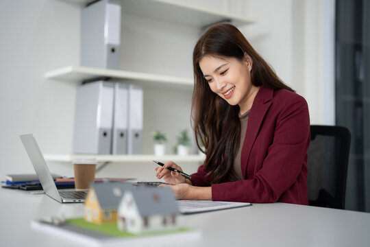 Asian professional real estate agent calculating property finances at modern office desk with house models for clients