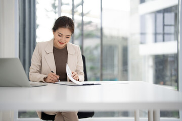 Asian adult woman real estate agent diligently reviewing sales documents in modern bright office workspace for client property deals