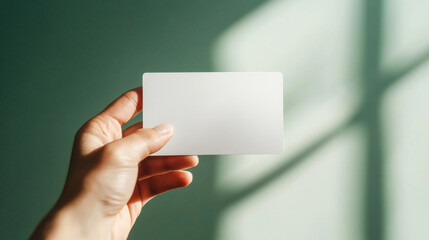 Human hand showing a minimalist blank white plastic card against a teal background with natural sunlight and shadows