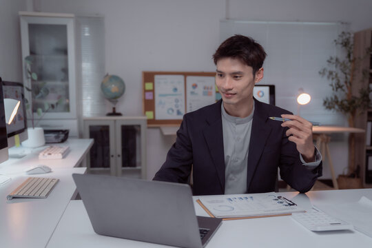 Young Asian businessman working late in a bright home office, focused on a video conference at his laptop while holding a pen and discussing important business matters