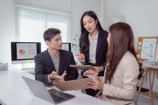 Asian business professionals collaborating on a project, discussing important data and financial charts with laptops and clipboards during a productive office meeting