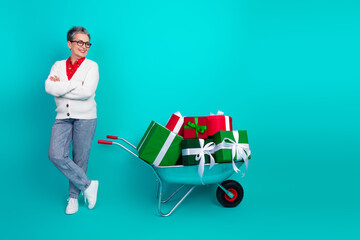 Joyful elderly woman with a wheelbarrow full of gifts in a teal studio ready for holiday shopping...