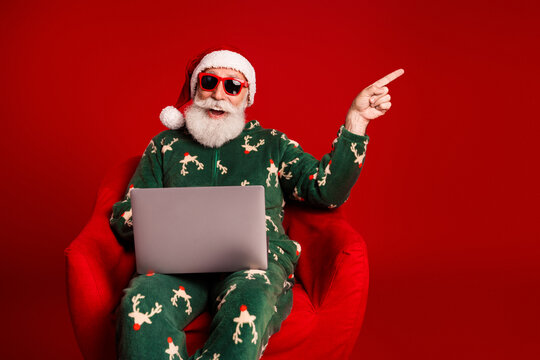 Santa in festive pajama sits in a red chair with a laptop wearing sunglasses and points to celebrate Christmas Eve from a warm home setting - Powered by Adobe