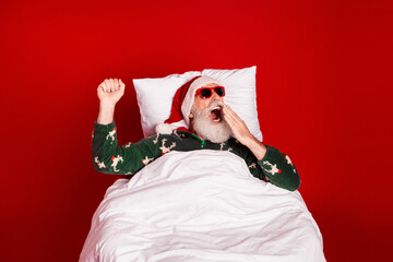 Naklejka na ściany i meble Santa in bed wearing red hat and green pajamas yawning with a cheerful festive vibe against a bold red background for christmas