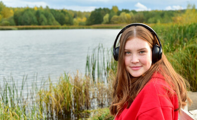 Young girl with long red hair outdoors, listening to music. Beautiful teenager girl in autumn park.
