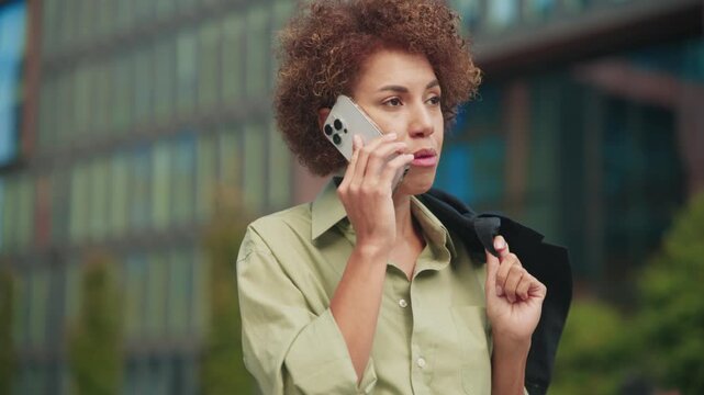 Focused African American woman with curly hair having serious conversation on smartphone. Wearing light green shirt and holding jacket on shoulder with one hand. Standing near office building outdoors