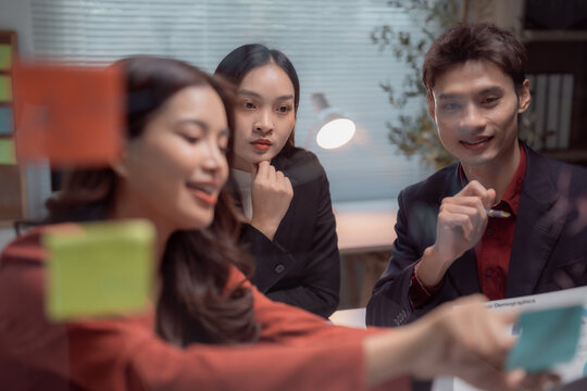 Asian business professionals collaborating and brainstorming ideas, working together in a corporate modern office, placing sticky notes on a glass board during a strategic planning meeting