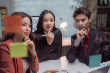 Three Asian business professionals discussing ideas and strategy, using sticky notes on a glass board during a collaborative brainstorming session in a modern office