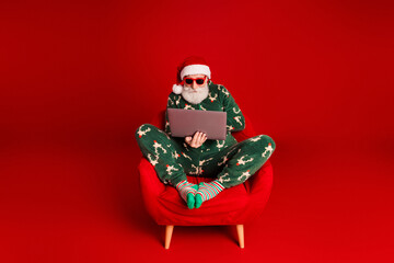 Naklejka na ściany i meble Santa in green pajama sits in a red chair using a laptop for Christmas online shopping and holiday fun in a bright red studio with merry festive vibes Naklejka na ściany i meble Santa in green pajama sits in a red chair using a laptop for Christmas online shopping and holiday fun in a bright red studio with merry festive vibes