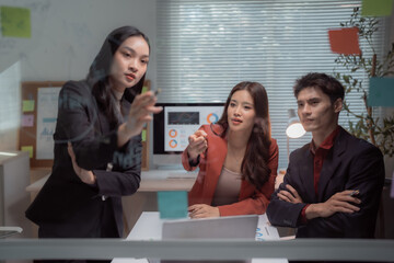 Young Asian business team collaborating and analyzing business data on a clear glass board in a modern office, developing strategies for project planning during a meeting
