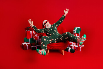 Naklejka na ściany i meble Santa in festive pajama lounge with gifts and red backdrop enjoying a cozy holiday moment for family and friends