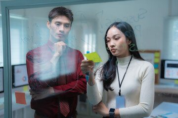 Young Asian business professionals collaborating in a modern office, planning strategy and writing ideas on a transparent glass board with sticky notes, symbolizing teamwork and innovation
