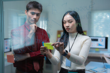 Asian business professionals collaborating on a strategic plan, writing and organizing sticky notes on a clear whiteboard during an important office meeting