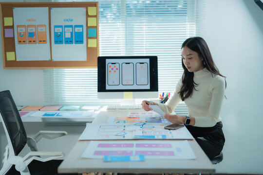 Woman analyzing mobile app wireframes and UI layouts at a desk, surrounded by paper mockups and a computer showing design iterations, focused on improving user experience