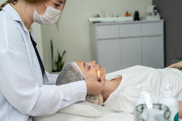 Naklejka premium Cosmetologist applies yellow gold alginate mask with spatula on woman face as anti-aging treatment