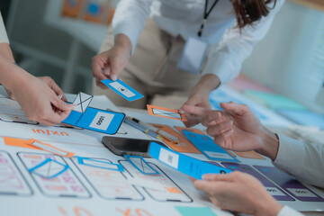 Designers' hands arranging paper components with mobile app interfaces, creating wireframes and mockups for user experience, engaging in a focused teamwork session