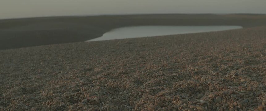 Real-time low-angle shot of the Suffolk shingle beach with a single still pool of water reflecting the morning sky, no sun or sea visible.