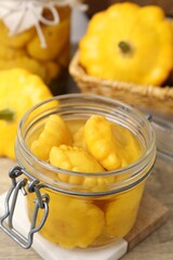 Pickled pattypan squashes in glass jar and fresh vegetables on wooden table, closeup