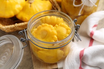 Pickled pattypan squashes in glass jars and fresh vegetables on wooden table, closeup