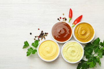 Different tasty sauces in bowls, parsley, peppercorns and chili peppers on white wooden table, flat lay. Space for text