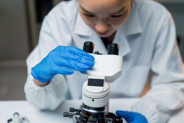 Asian woman scientist wearing lab coat and blue gloves, looking into microscope, conducting...