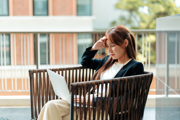 Fototapeta premium Asian businesswoman suffering from severe headache and stress, holding her head while working on a laptop remotely