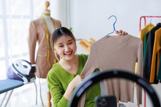 Smiling Asian woman presenting clothing on live stream, promoting small business products on social media
