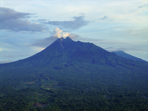 Aerial view of a lush, green volcanic mountain rising majestically, with a plume of smoke against the blue sky, Merapi Mountain, Klaten, Central Java, Indonesia.