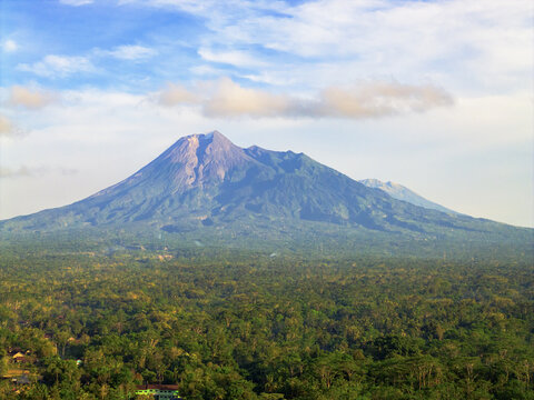 Aerial view of imposing Merapi Mountain rises majestically, its slopes a tapestry of grey rock and verdant green, under a sky brushed with wisps of clouds, Klaten, Central Java, Indonesia.