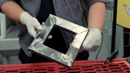 Metalworker holding welded steel frame piece. A worker wearing gloves shows a freshly welded square steel frame with visible weld marks, inside a metal workshop environment.