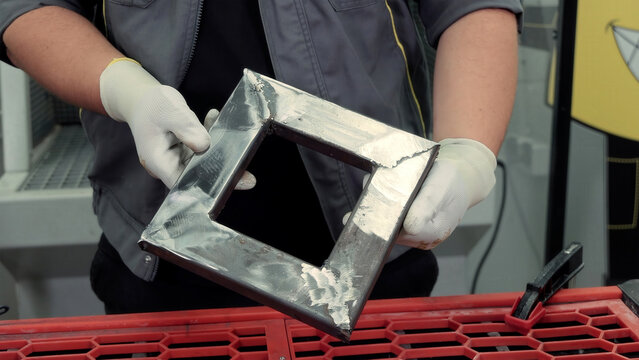 Metalworker holding welded steel frame piece. A worker wearing gloves shows a freshly welded square steel frame with visible weld marks, inside a metal workshop environment.