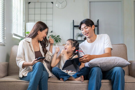 Asian family sitting on sofa, parents and child looking happy while giving thumbs up, using mobile phone and tablet - Powered by Adobe