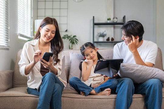 Asian family relaxed on sofa, parents and daughter sharing tablet while mother scrolls smartphone, smiling and bonding at home - Powered by Adobe