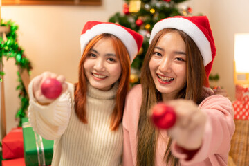 Two Young Adult Women Celebrating Christmas Holiday Season with Red Ornament Balls and Santa Hats near Christmas Tree and Gifts at Home Party