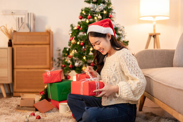 Young Woman Opening Christmas Present With Santa Hat and Christmas Tree Sparkly Lights Indoors Holiday Joy Celebrating Merry Xmas Gift Giving Season